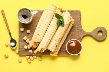 Board with delicious wafer rolls, boiled condensed milk, cup of coffee and hazelnuts on yellow background