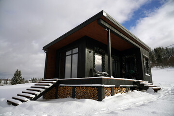 Modern black tiny cabin in snowy weather with stacked firewood under