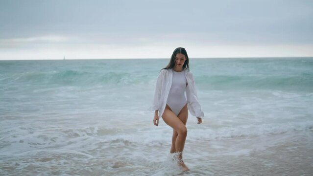 Woman walking sea foam in sexy white swimsuit. Young girl going on ocean beach.