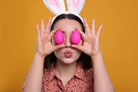 Happy woman in bunny ears headband holding painted Easter eggs near her eyes on orange background