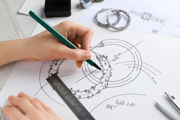 Jeweler drawing sketch of elegant earrings on paper at white wooden table, closeup
