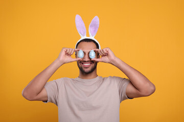 Happy African American man in bunny ears headband covering eyes with Easter eggs on orange background