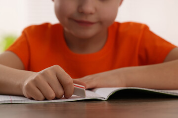 Little boy erasing mistake in his notebook at wooden desk, closeup