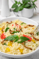 Bowl of delicious couscous with vegetables and basil served on white wooden table, closeup