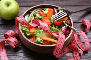 Bowl with fresh vegetable salad, fork, apple and measuring tape on wooden table, closeup. Healthy diet concept