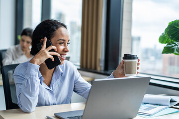 Latino happy businesswoman talking on smartphone and working in office. Attractive young girl employee worker in formal wear sitting on table and use computer plan work project at corporate workplace.