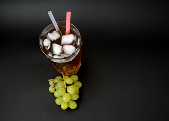 A tall glass of fruit juice with ice and straws on a black background, next to a bunch of ripe green grapes.