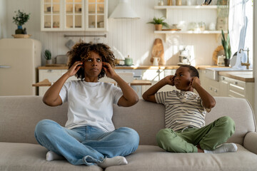 Unhappy irritated African American family mother and son sitting on sofa covering ears annoyed by...