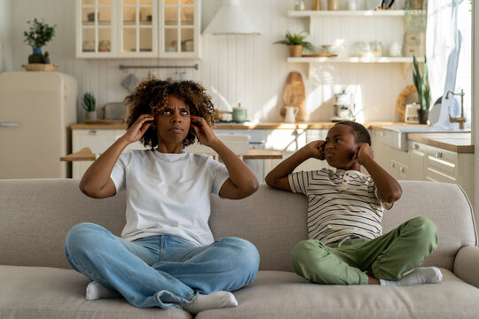 Unhappy Irritated African American Family Mother And Son Sitting On Sofa Covering Ears Annoyed By Loud Noisy Neighbors In Poorly Insulated Apartment, Living In House With Poor Sound Insulation