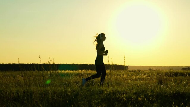Training run with music. Running after sun. Healthy girl listen to music in headphones is engaged in fitness, runs in meadow, sun. Girl runner athlete outdoors. Young woman runs in park at sunset