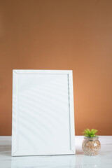 Blank white frame mockup placed on wooden table and flower vase. Elegant workspace on white brown background.