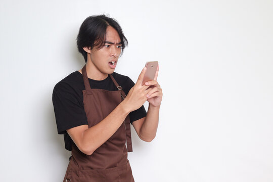 Portrait of shocked Asian barista man in brown apron looking at his mobile phone with surprised expression. Advertising concept. Isolated image on white background