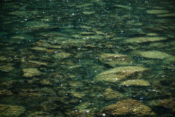 Meditative ripple and many reflected lights in green mountain lake. Beautiful relaxing background of stony bottom in turquoise transparent water of glacial lake in sunlight. Many stones under water.