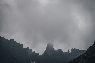 Dark atmospheric mountain landscape with black pointy rocky peak in gray cloudy sky. Lead gray low clouds among black mountains. Dark rocky pinnacle in low clouds in rainy weather. Gloomy minimalism.