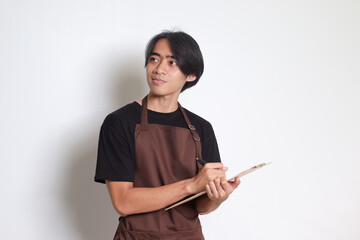 Portrait of attractive Asian barista man in brown apron taking order, writing on menu book list. Isolated image on white background