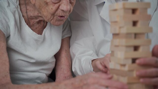 Jenga game. Theme is dementia, aging and games for old people. Caucasian senior woman builds tower of wooden blocks with the help of a doctor as part of a therapy and jenga game at a patient's home.