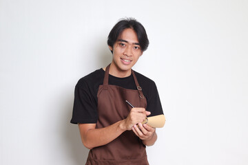 Portrait of attractive Asian barista man in brown apron taking order, writing on menu book list....