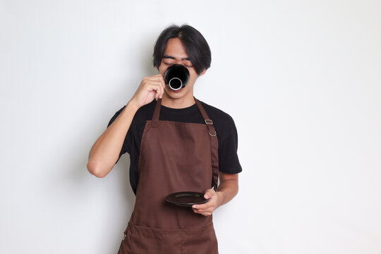 Portrait of attractive Asian barista man in brown apron drinking a cup of coffee while holding the saucer. Isolated image on white background