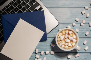 Empty greeting or invitation card mock up blue envelope with white cup of coffee and marshmallows laptop keyboard on wooden blue table. Workplace Blank paper copy space for your text. Valentines day