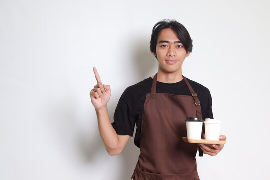 Portrait of attractive Asian barista man in brown apron holding take away paper coffee cup with wooden tray to serve while pointing to the side. Isolated image on white background
