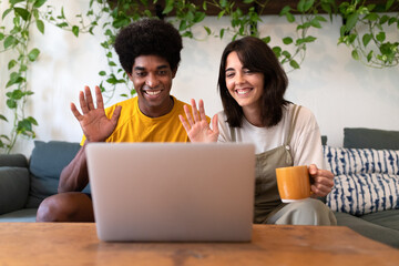 Young multiracial couple sitting on sofa waving hello on a video call using laptop.