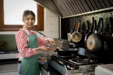 Beautiful asian girl doing a stir-fry gesture on the pan while standing and looking to the camera with smile on her face in kitchen backgound.