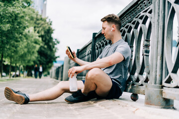 Fototapeta na wymiar Tired Young man runner sitting on road, using mobile phone and relaxing after sport training. Holding water bottle while doing fitness workout in city urban street, cloudy sky at summer.