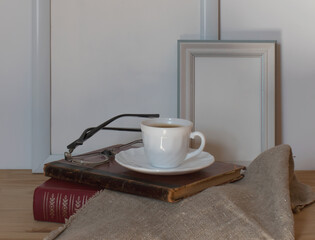 A white porcelain cup and saucer with black coffee, glasses on a stack of old books against the background of empty white frames for photos or paintings.