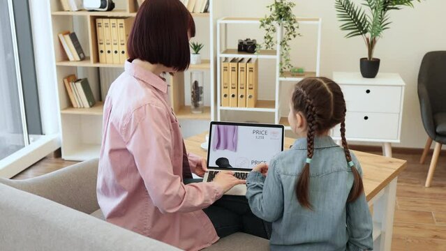 Rear view of dark haired woman sitting on couch with laptop on knees while her pretty girl pointing on screen with forefinger. Mother and daughter choosing new clothes to buy during discounts.