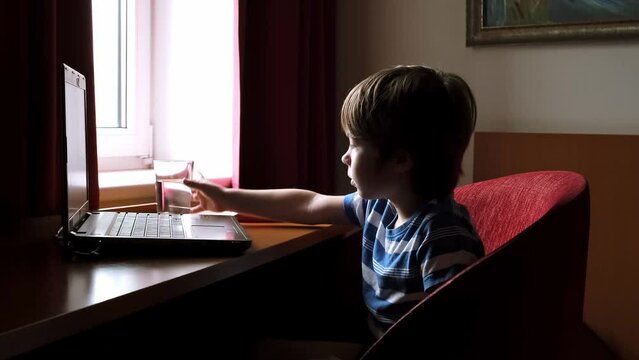 6 year boy sitting at a table carefully looking into a laptop screen and drinking water from glass beaker. Child watching cartoons. Healthy habit. Water balance. High speed internet. Online education.
