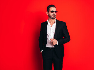 Obraz na płótnie Canvas Portrait of handsome confident stylish hipster lambersexual model. Sexy modern man dressed in black elegant suit. Fashion male posing in studio, isolated on red. In sunglasses