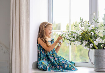 Cute baby girl Caucasian in a green floral sundress sitting on the windowsill by the window. Decor. Home comfort. Spring concept.