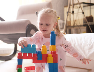 Naklejka premium A laughing little child, a blonde preschooler playing with colorful cubes, sitting on the floor in pink pajamas, in a sunny room with a large window at home. Early development of children.