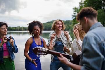 A group of young friends having fun near a lake, laughing and playing guitar.
