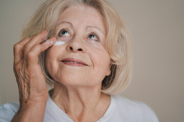 Smiling old caucasian woman applying anti aging cream on her face against grey background. Beauty...