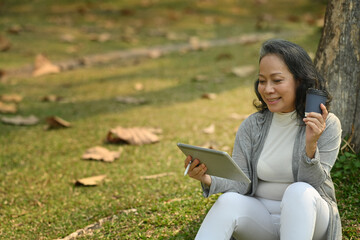 Positive middle aged woman holding paper cup of coffee using digital tablet under tree in the park at sunny beautiful day