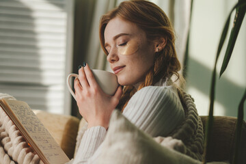 Attractive woman sitting on sofa in the morning, drinking tea, reading book, feeling comfortable at...