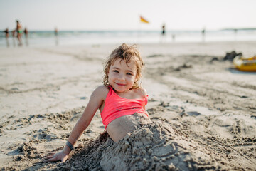 Little girl buring herself in sand, enoying summer vacation at sea.