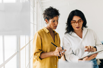 Two business women having a discussion, they're standing in an office and using a tablet