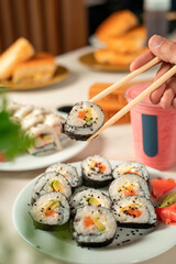 Chopsticks holding a fresh sushi maki with wasabi and ginger in wooden table with drinks