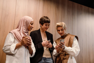 Group of cheerful female colleagues using mobile phone and drinking coffee during meeting