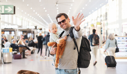 Father traveling with child, holding his infant baby boy at airport terminal waiting to board a plane waving goodby. Travel with kids concept