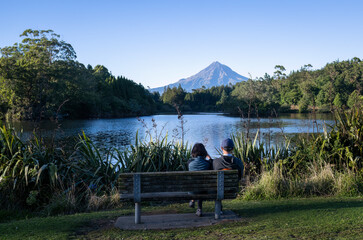 Fototapeta na wymiar Couple sitting on the bench and enjoying the view of Mt Taranaki at Lake Mangamahoe. New Plymouth.