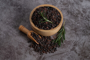 Himalayan black salt, Indian black salt or Kala namak in wooden bowl with wooden scoop and rosemary on black stone table. Used in cooking, especially in Asian and Indian cuisine. Bath salt spa.