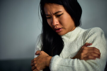 Close-up of an anxious Asian woman, looking down while sitting on a couch.