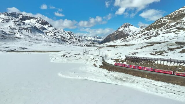 Long red train driving over a railway track in a winters landscape in Switzerland. Drone panning shot