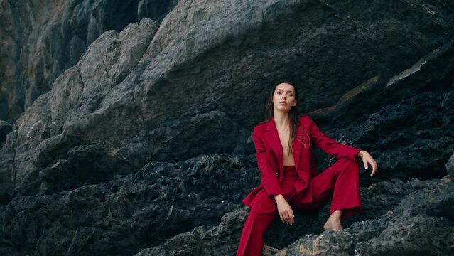 Confident woman sitting mountain ledge in red suit. Model posing at stone rock.