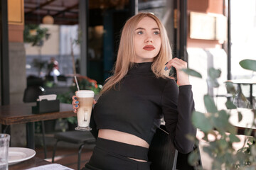 Brooding attractive young blonde woman sits at table in coffee shop with cup of cappuccino coffee latte.