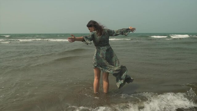 Girl with flowing long dress slowly turning with waves breaking around her. Cinematic slow mo video.