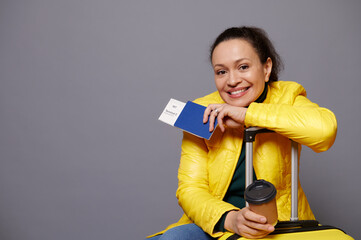 Attractive cheerful Latin American woman passenger, smiling broadly looking at camera, posing with yellow suitcase, air flight ticket and takeaway coffee in eco cardboard cup, gray isolated background